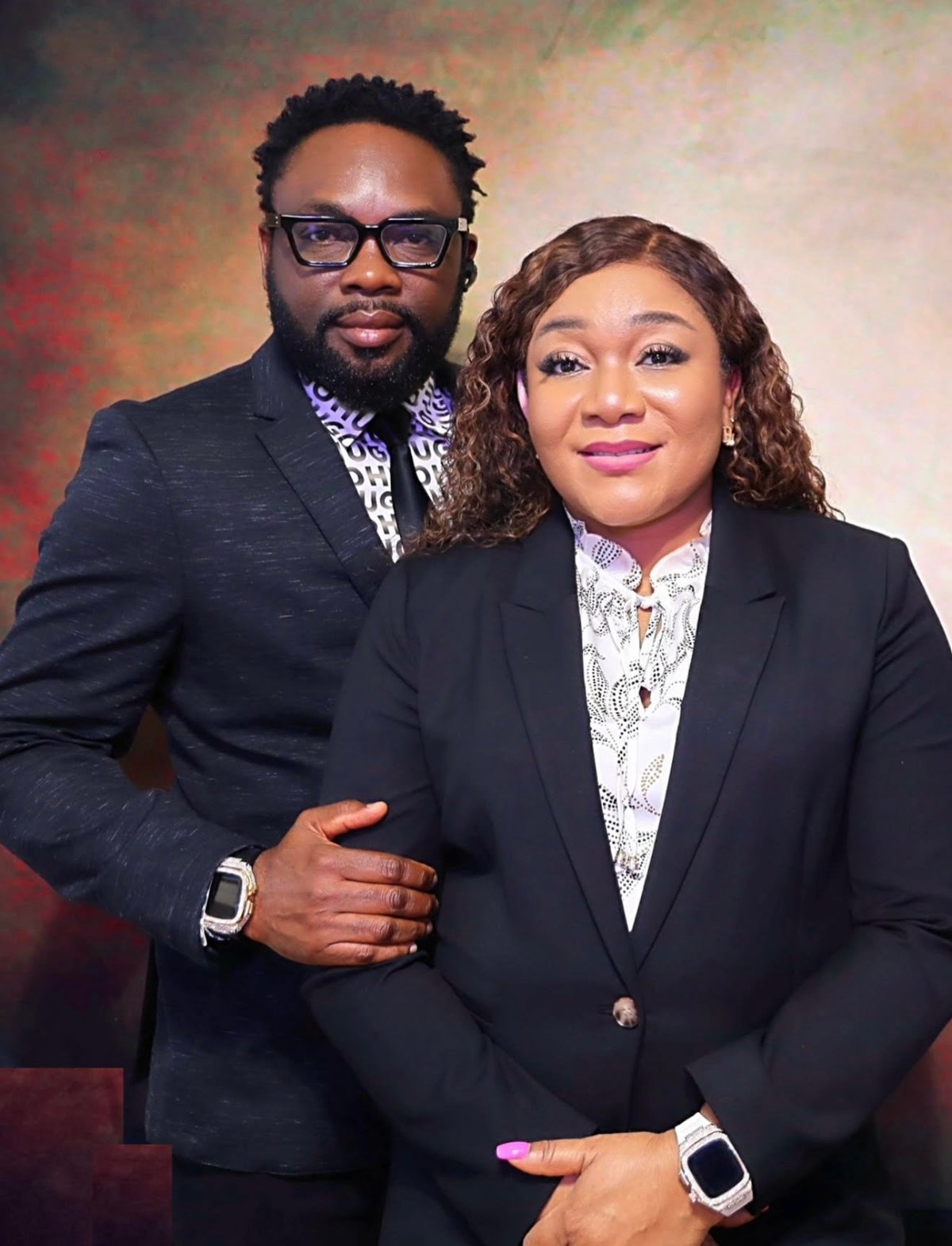 Two professionals in formal black blazers pose together against a textured red and beige backdrop for a portrait photo